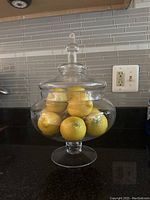 Front view of apothecary-style clear glass pedestal covered vase filled with several round yellow faux lemons on a dark countertop against a tiled backsplash.