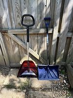 Two snow shovels placed side by side against a wooden gate, one red scoop shovel and one purple scoop shovel, showing the handles and blades.