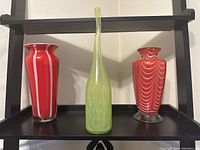 Photo showing three decorative vases on a black shelf; two pink with white stripes and one tall lime green vase.