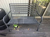 Black wrought iron bench with horizontal slats and armrests, shown on wooden deck outdoors with background items including stacked tires and garden decoration.