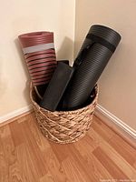 Photo of a woven basket containing two rolled yoga mats—one black and one reddish with stripes—two black yoga bricks, and other small items inside the basket.