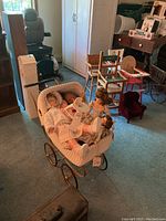 Full view of collector dolls seated in vintage white doll carriage inside room with various chairs and other furnishings
