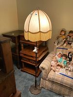 Full view of the floor lamp showing metal base, tall slender pole, and fabric shade with tassels around edge. Lamp is placed next to a couch and wooden side tables.