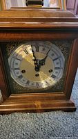 Close-up of Howard Miller mantle clock face showing Roman numerals and ornate black hands with brass and wooden framing.