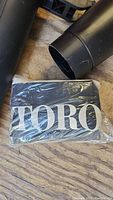 Toro branded vacuum bag wrapped in plastic on wooden floor with blower vac tubes nearby