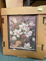 Framed wall art featuring white flowers and fruit wrapped in plastic inside a cardboard box
