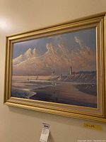 Framed coastal scene with seagulls flying over the ocean near a lighthouse under a cloudy sky, gold frame, signed by August Holland, measuring 20x28.