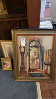 Framed artwork showing an interior entrance hall with a grandfather clock, an arched window view of a garden, and floral arrangement on a side table.