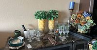 Display of two yellow vases with artificial greenery, various clear stemware, and one bronze candle holder with blue candle on buffet table