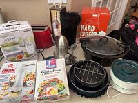 Photo showing various small kitchen appliances and boxes including a NutriChef electric food steamer, red toaster, cookie press sets, a juicer and large cooking pot with lid.