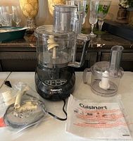 Photo shows the full Cuisinart food processor with large clear bowl on the base, accessory pieces in a bag, and manual on the table.