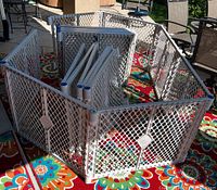 Grey plastic mesh playpen panels assembled in an octagon shape outdoors on a multicolor patterned rug; additional panels stacked inside.