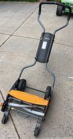 Full image of a manual push reel lawn mower with orange and black body and a metal handle, showing front view with plastic wheels on concrete surface.