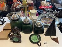 Green glass canister set with lids, decorative metal lantern with green glass, and pyramid-shaped black metal wind chime with chain and triangular metal clapper all arranged on table.