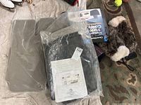 Photo showing two new GMC rubber car mats in original plastic packaging, a boxed snow trunk caddy organizer, and two fur-lined snow scrapers pushed on a floor mat surface in a garage.