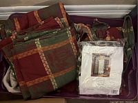 Top view of folded harvest theme table linens including tablecloths and placemats in deep red, green, and orange patchwork with fall motifs inside a purple plastic bin.