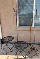 Photo showing two black metal mesh patio side tables with foldable legs, one black metal hanging plant hook stand with multiple hooks and tripod base, and one taller bronze-colored metal hanging plant hook stand with single hook and tripod base, all weathered.