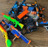 Top view containing multiple Nerf dart blasters in orange, blue, green, and yellow colors, a Nerf vest and orange blue foam darts and orange clip.