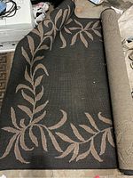 Rolled outdoor rug showing black base with beige leaf pattern design and woven texture