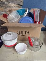 Box filled with various plastic food storage containers with lids, including red, blue, and clear plastic pieces, small white ramekin and glass Pyrex pieces outside the box.
