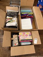 Three open boxes filled with various school and office supplies including notebooks, folders, and lined paper.