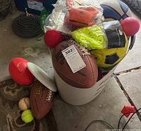Basket filled with assorted sports balls and accessories including footballs, soccer ball, tennis balls, baseball, flyer disc, all on a carpeted floor.