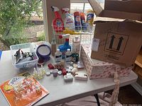Wide view of assorted cleaning sprays, toiletries, and miscellaneous items on table with open cardboard box