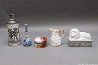 Front view of 5 items including German beer stein, Delft vase, trinket box, Royal Stafford pitcher, and white lion figurine on decorated base arranged on gray background
