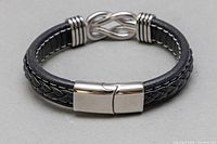 Front view of black leather bracelet with braided and smooth bands, silver-tone infinity symbol in center, and polished silver clasp.