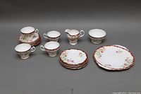 Full set of bone china: 4 teacups on 6 saucers at left, 6 dessert plates stacked at center back, creamer and sugar bowl to right, and a large 10-inch cake plate in front right. Floral pattern with maroon rim and gilded edges visible.