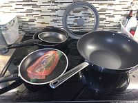 Four cooking pans displayed on a stove including frying pans, sauté, and wok style pan, one glass lid visible.
