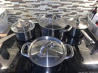 Four stainless steel pots with glass lids arranged on stovetop, showing handles and lid details.