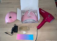 Photo of two pink nail lamps (one in box), Conair hair dryer, exfoliating brush, and two nail files on wooden surface.