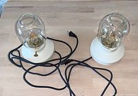 Two white-based bedside lamps with clear globe shades placed side by side showing power cords and switches.