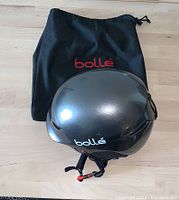 Metallic gray Bolle helmet shown on top of the black Bolle branded fabric drawstring carry bag on a wooden surface.