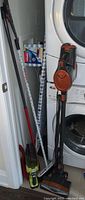 Full view of all cleaning supplies including Rocket upright vacuum, Rioby handheld vacuum, ironing board, broom, and Vileda mop in a laundry room.