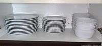 Two stacks of white ceramic plates on the left and medium white ceramic bowls on the right on a shelf
