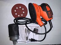 Black & Decker orange and black random orbit sander with attached power cord, dust collection bag, sanding disc, and manual on flat surface.