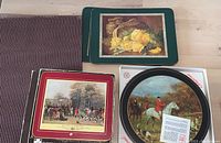Photo showing three rectangular placemats with floral and hunting-themed artwork with green, red, and black borders