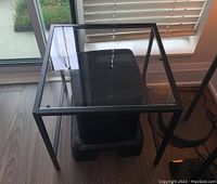 Square glass top side table with black metal frame shown near a window with a visible floor lamp and black basket beneath.