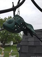 Side view of a greenish cast iron fish hanging outdoors from a metal support arm, showing open lattice body and detailed fins and eyes.