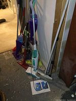 View of multiple cleaning tools leaning against wall on floor including Shark steam mop, Swiffer mop, Bissell sweeper and dusters.