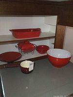 Red rectangular lasagna pan, casserole dish with warming stand, red serving bowls, and a small red sauce pot with cream interior and handle on countertop and shelves.