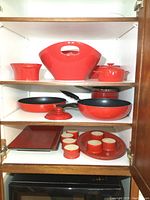 Cabinet shelf with large red serving bowl, covered casserole dish, two frying pans with lids, round serving tray with six ramekins, and rectangular platter.