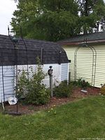 Wide view showing the pair of black metal garden arbours set near garden beds by a shed and house, displaying overall size and placement.