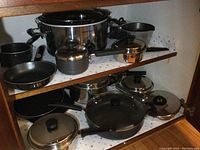 View of cupboard shelf showing Hamilton Beach slow cooker, several stainless steel pots and pans, some with black handles and lids.
