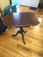 Side view of small wooden drop leaf side table showing the extended leaf with scalloped edges and the turned pedestal leg with claw feet on hardwood floor.