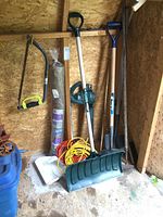 Photo of assorted yard tools including green snow shovel, electric leaf blower, extension cords, hand shovels and more inside a wooden shed.