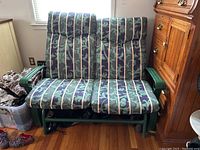 Outdoor glider bench resting indoors on wooden floor near wooden cabinet. Cushions are installed and bench is visible from front.