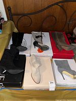 Overview of all six pairs of boots arranged on boxes, showing variety in colors including black, gray, beige, and taupe, with different heel types and heights.